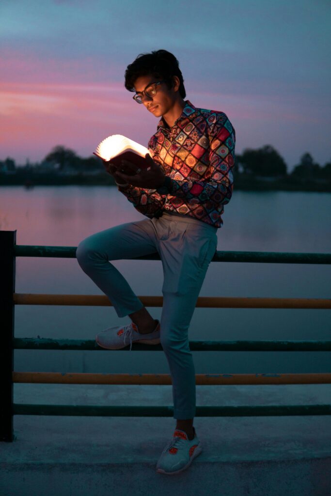 A young man reads by a lake at sunset, enjoying a moment of leisure and relaxation.